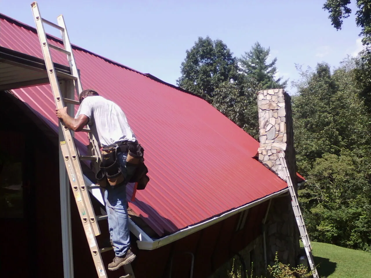 Roofer on a ladder beside a red metal roof and stone chimney — Soffit & Fascia Repair by Roofing Brothers in Canal Winchester
